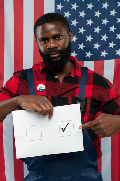 Jeune Réparateur Afro-américain Contemporain Pointant Sur Tick Dans L'un Des Carrés Sur Le Bulletin De Vote En Se Tenant Debout Contre Le Drapeau Américain