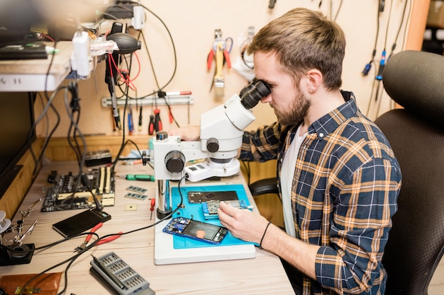 Jeune réparateur barbu assis par table en face du microscope pour voir de petits détails de gadgets cassés en atelier