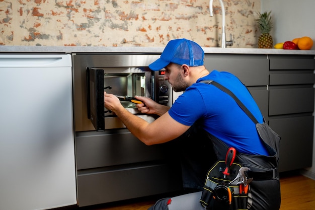 Jeune réparateur en uniforme bleu répare et répare un four à micro-ondes avec un tournevis dans une cuisine moderne