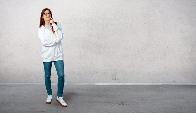 Jeune rousse dans un sweat-shirt blanc urbain avec des lunettes debout et pensant