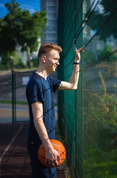 Un jeune roux vêtu d'un t-shirt bleu foncé se tient avec un ballon de basket à la main sur le terrain de sport avec un anneau de boxetball sur fond de maille