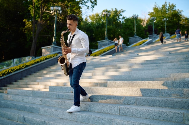 Le jeune saxophoniste joue du saxophone dans les escaliers dans le parc d'été. Musicien avec saxophone à l'extérieur, performance musicale dans la nature, interprète de mélodie de jazz