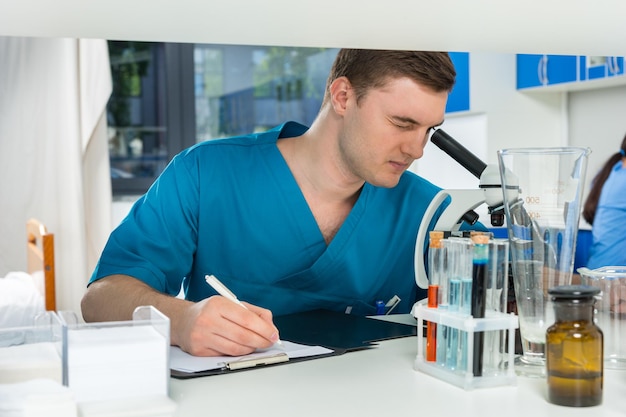 Un jeune scientifique en uniforme regarde à travers un microscope et va prendre des notes sur ses recherches dans un laboratoire. Concept de soins de santé et de biotechnologie