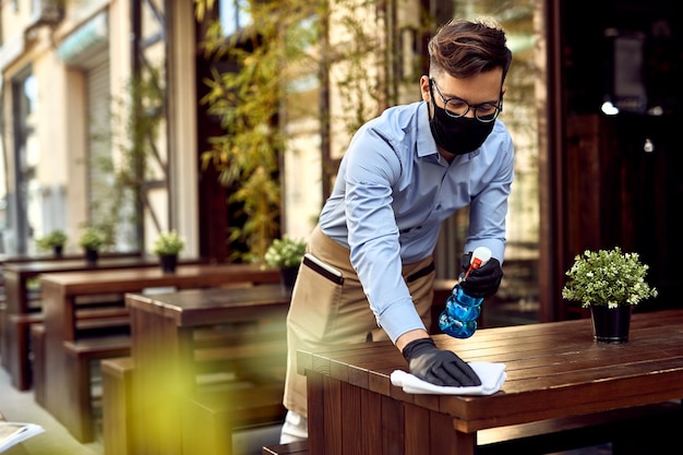 Jeune serveur nettoyant des tables au café en plein air après sa réouverture après l'épidémie de coronavirus