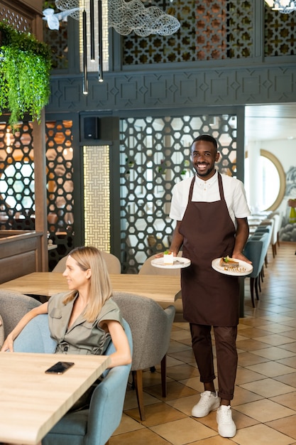 Jeune serveur souriant portant des assiettes avec des commandes de femmes détendues assises dans des fauteuils à table et parlant au café