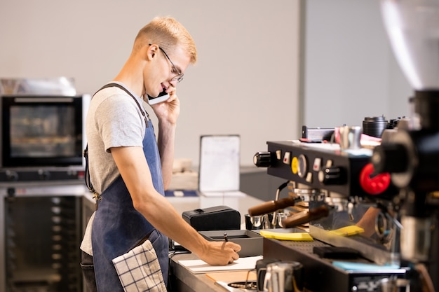 Jeune serveur ou travailleur de la cafétéria en écrivant les commandes des clients dans le bloc-notes tout en leur parlant sur téléphone mobile par lieu de travail