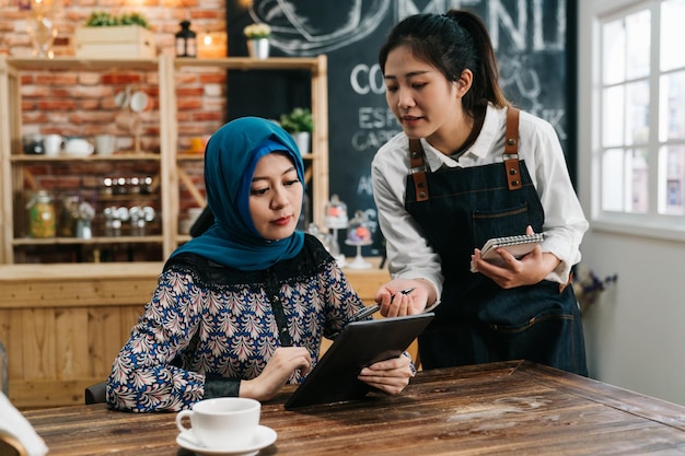 Jeune serveuse asiatique conseillant à une cliente islamique de choisir une nouvelle collation dans le menu sur un bloc-notes mobile au restaurant. personnel du café fille prenant la commande d'une cliente musulmane au café-bar pour le déjeuner.