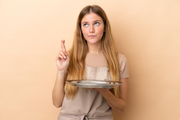 Jeune serveuse blonde femme avec plateau isolé sur fond beige avec les doigts croisés et souhaitant le meilleur