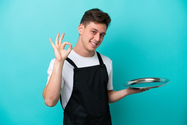 Jeune serveuse avec plateau isolé sur fond bleu montrant le signe ok avec les doigts