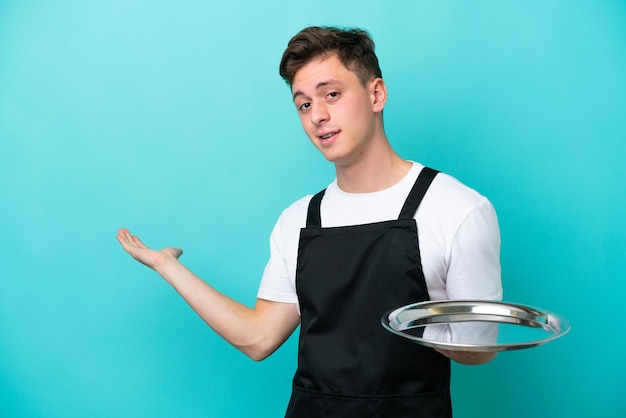 Jeune serveuse avec plateau isolé sur fond bleu tendant les mains sur le côté pour inviter à venir