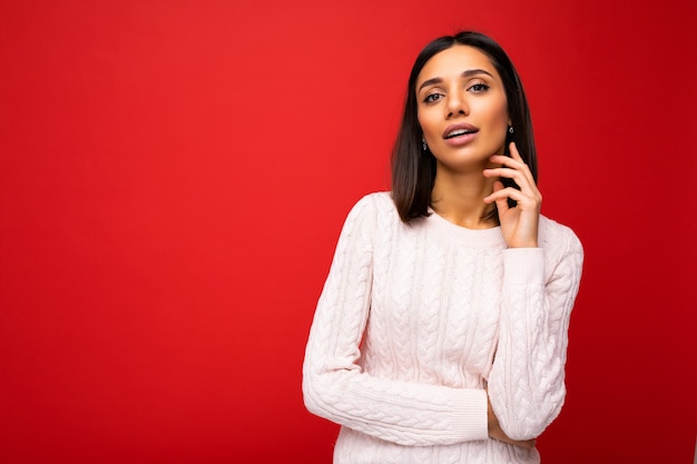 Jeune sexy charmante belle femme brune avec des émotions sincères portant un pull décontracté isolé sur un mur rouge avec espace de copie.