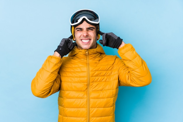 Jeune skieur homme portant des lunettes de snowboard couvrant les oreilles avec les mains.