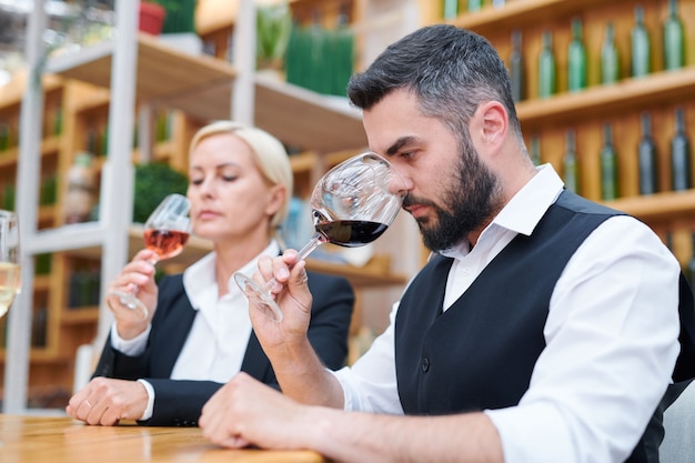 Jeune sommelier barbu en tenue de soirée sentant une nouvelle sorte de vin rouge dans un verre à vin tout en examinant sa qualité