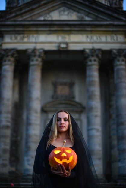 Une jeune sorcière sinistre tient une citrouille avec un visage terrible contre le château.