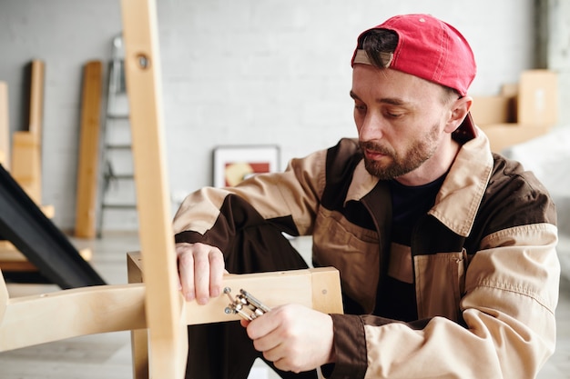 Jeune spécialiste sérieux de l'assemblage de meubles dans les vêtements de travail fixant des pièces de chaise tout en l'assemblant dans une grande maison neuve, un appartement ou un studio