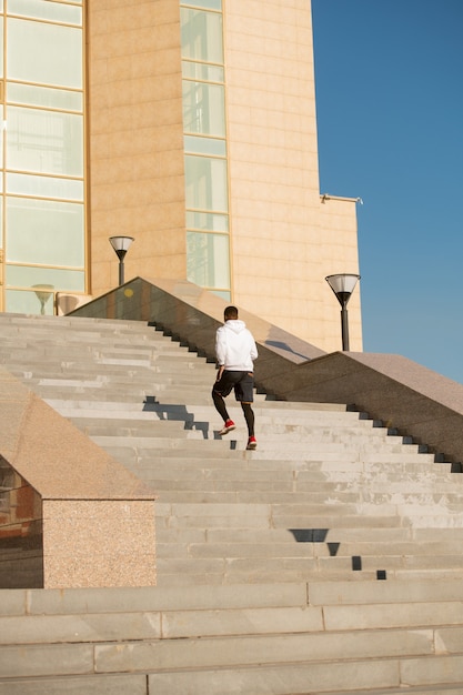 Jeune sportif africain en vêtements de sport assis sur des balustrades en marbre par un grand escalier devant l'architecture moderne en milieu urbain