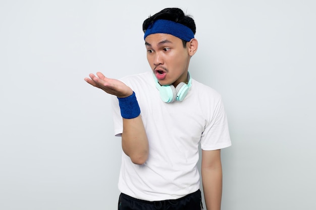 Jeune sportif asiatique en bandeau bleu et t-shirt blanc sportswear avec un casque confus pour voir quelque chose dans la paume isolé sur fond blanc Concept de sport d'entraînement