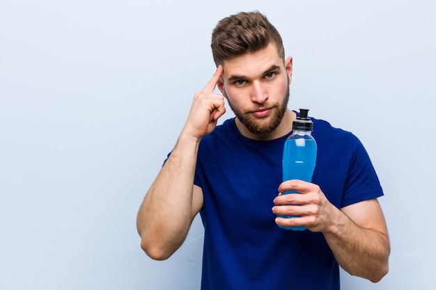 Jeune sportif caucasien tenant une boisson isotonique pointant sa tempe avec le doigt, pensant, concentré sur une tâche.