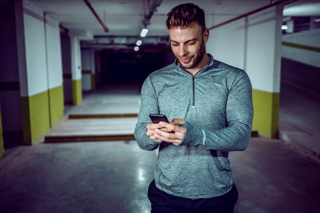 Jeune sportif caucasien en vêtements de sport à l'aide d'un téléphone intelligent et se vanter de sa réussite. Garage souterrain. La nuit.