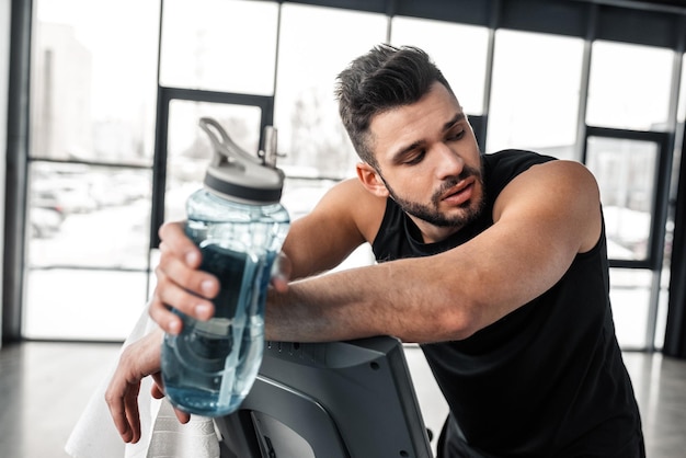 Jeune sportif fatigué se penchant sur le tapis roulant et tenant une bouteille de sport avec de l'eau dans la salle de sport