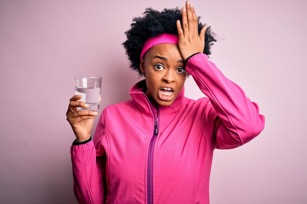 Jeune sportive afro-américaine aux cheveux bouclés buvant un verre d'eau stressée avec la main sur la tête choquée par la honte et la surprise face en colère et frustrée Peur et contrariée par une erreur