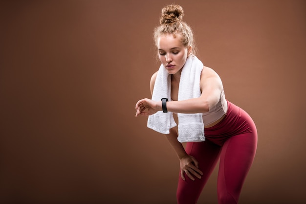 Jeune sportive blonde fatiguée en vêtements de sport regardant fitbit sur son poignet tout en faisant un exercice physique difficile sur brown