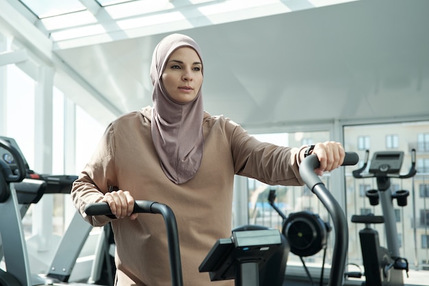 Jeune sportive en hijab tenant par des poignées d'équipements sportifs tout en faisant de l'exercice dans un centre de remise en forme moderne