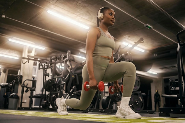 Une jeune sportive noire en forme s'entraîne à faire des sauts avec des haltères et à écouter de la musique.