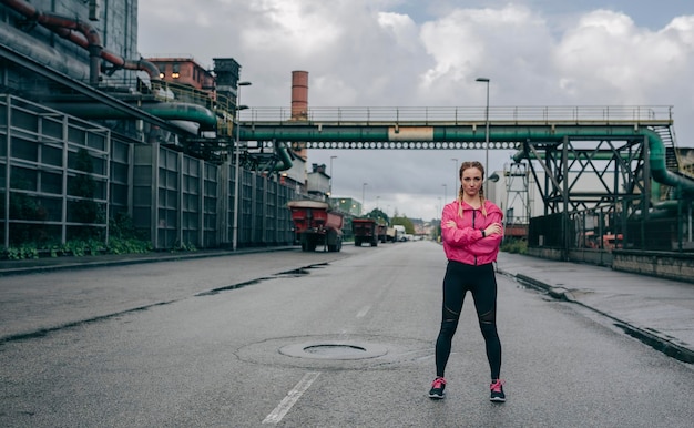 Jeune sportive posant les bras croisés à l'extérieur un jour de pluie dans une zone industrielle