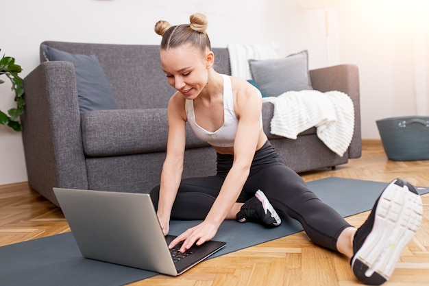 Jeune sportive en sportswear souriant et parcourant un ordinateur portable alors qu'il était assis sur un tapis pendant l'entraînement en ligne à la maison