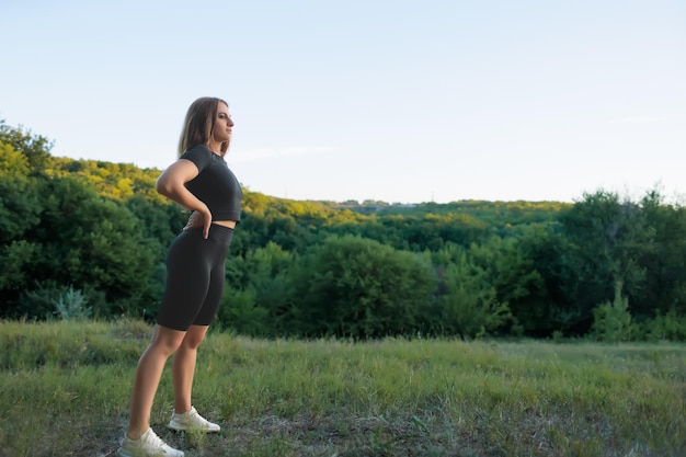 Une jeune sportive en survêtement noir garde ses mains sur sa ceinture et regarde le coucher de soleil dans le parc