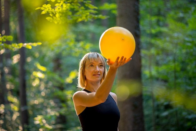 Jeune stagiaire blonde en pilates tenant une balle jaune tout en s'entraînant dans la nature.