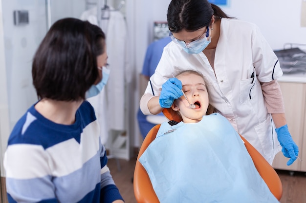 Jeune stomatologue utilisant un miroir incurvé pour vérifier le spécialiste de la dentisterie lors de la consultation de la cavité de l'enfant dans un bureau de stomatologie utilisant la technologie moderne.