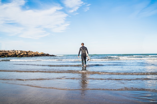 Jeune surfeur entrant dans l'eau avec sa planche de surf dans une combinaison de surf noire. Concept de sport et de sports nautiques.
