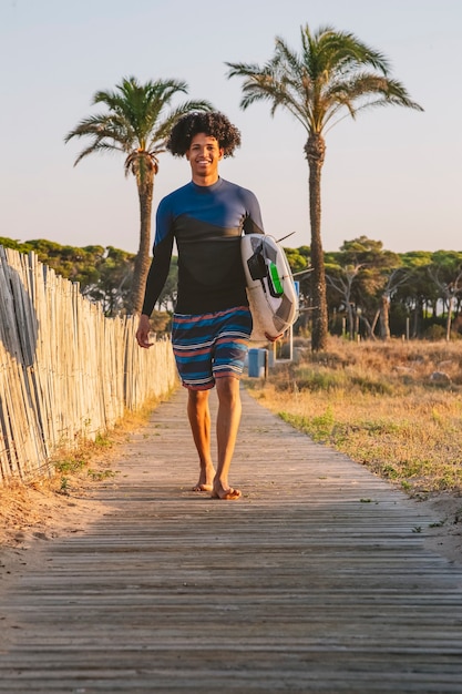 Un jeune surfeur latino afro descend la passerelle en bois jusqu'à la plage avec une planche de surf au lever du soleil