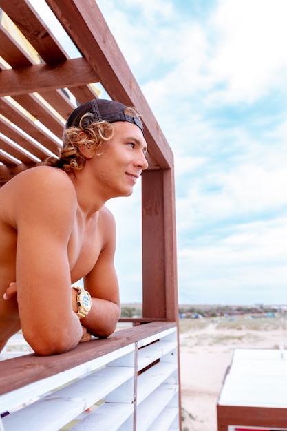 Photo jeune surfeur mec attrayant et souriant debout sur la terrasse d'une école de surf en regardant la distance