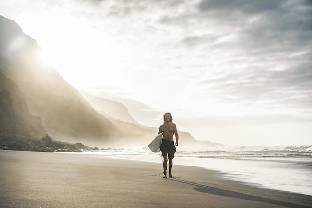 Jeune surfeur sur une plage tropicale au coucher du soleil - homme avec sa planche de surf marchant à côté de l'océan par une journée ensoleillée - concept de sport extrême - se concentrer sur le corps masculin