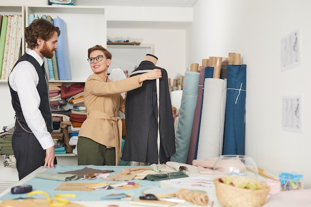 Jeune tailleur en lunettes mesurant le dos de la veste sur mannequin et discutant de quelque chose avec son client dans l'atelier