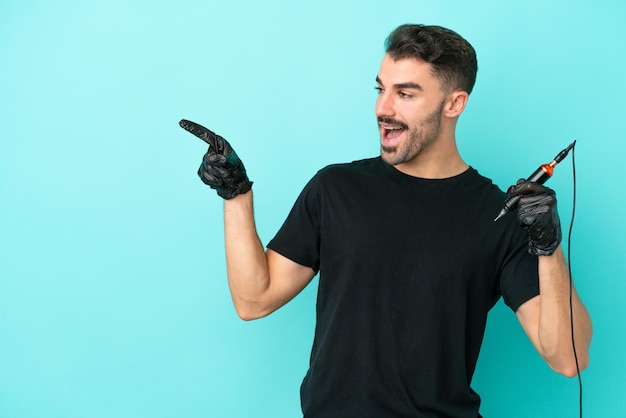 Jeune tatoueur homme isolé sur fond bleu pointant le doigt sur le côté et présentant un produit