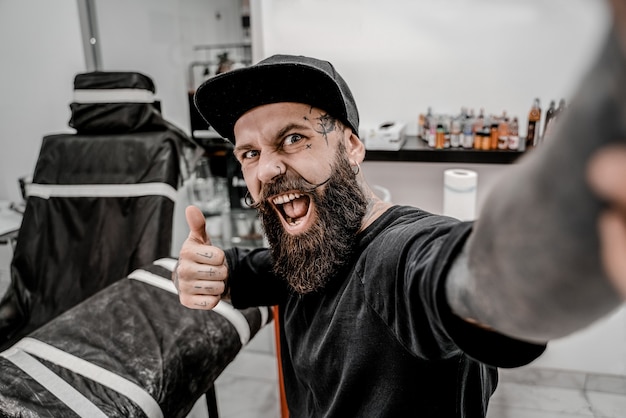 Jeune tatoueur masculin avec barbe prenant selfie et a l'air drôle dans le lieu de l'atelier avant le travail.