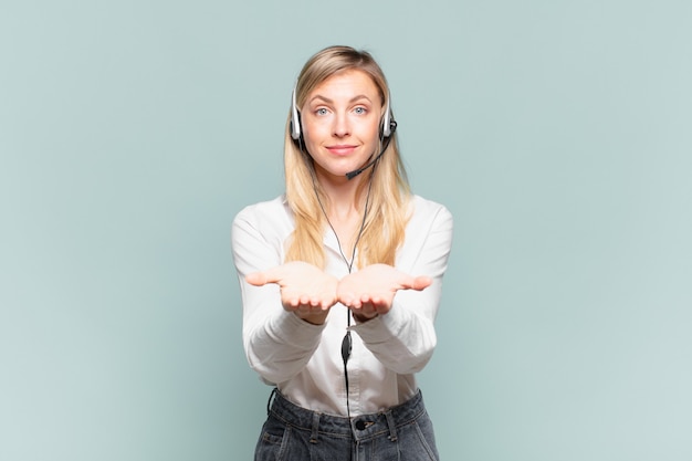 jeune télévendeuse blonde souriante joyeusement avec un regard amical, confiant et positif, offrant et montrant un objet ou un concept