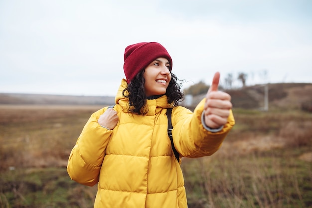 Jeune touriste attrape une voiture sur la route d'hiver loin de la ville. Concentrez-vous sur le visage. Femme portant une veste jaune, un sac à dos et un chapeau rouge.