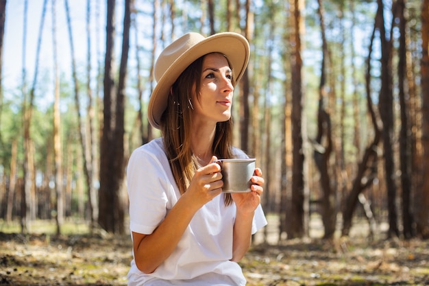 Jeune touriste en chapeau et t-shirt boit du thé ou de l'eau lors d'une halte dans la forêt.
