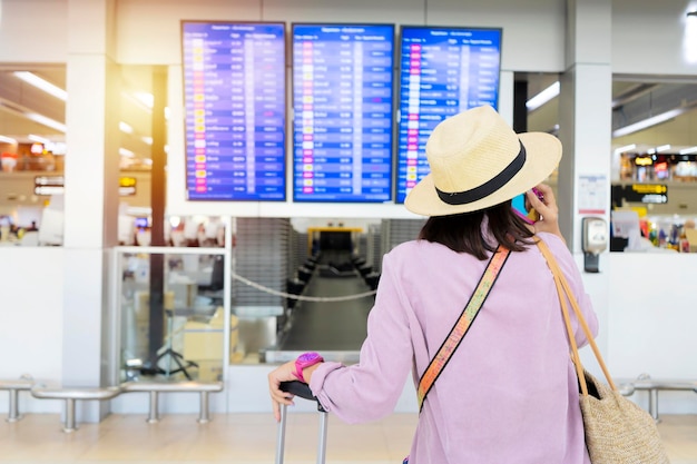 Jeune touriste élégant avec bagage à main dans le terminal de l'aéroport international regardant le panneau d'information vérifiant son vol Membre d'équipage de cabine avec valise