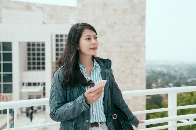 Jeune touriste élégante jolie femme utilisant l'application smartphone sur le bâtiment de la ville en plein air. voyageur coréen asiatique s'appuyant sur la balustrade tenant un téléphone portable et regardant la vue. dame routard se détendre profiter du tourisme