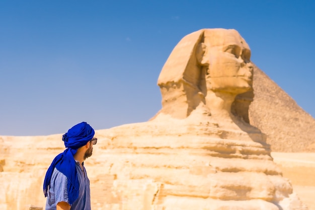 Un Jeune Touriste Près Du Grand Sphinx De Gizeh Vêtu De Bleu Et D'un Turban Bleu, D'où Les Miramides De Gizeh, Le Tourisme Culturel Et Beaucoup D'histoire. Le Caire, Egypte