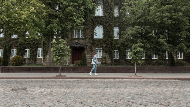 Un jeune touriste urbain élégant dans des vêtements de jeans pour jeunes à la mode avec un sac en tissu à la mode voyage dans une rue de la ville vintage près d'un vieux bâtiment envahi par les feuilles vertes. Le gars de la mode moderne aime marcher.