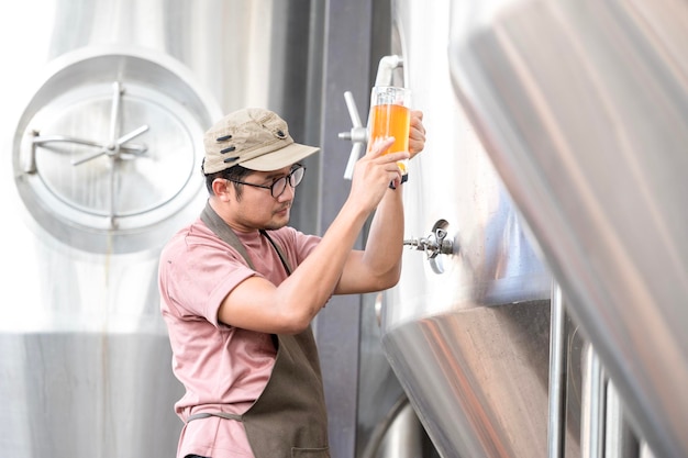 Jeune travailleur asiatique inspectant la qualité de la brasserie avec un verre de bière artisanale évaluant l'apparence visuelle après préparation tout en travaillant dans une brasserie artisanale de transformation