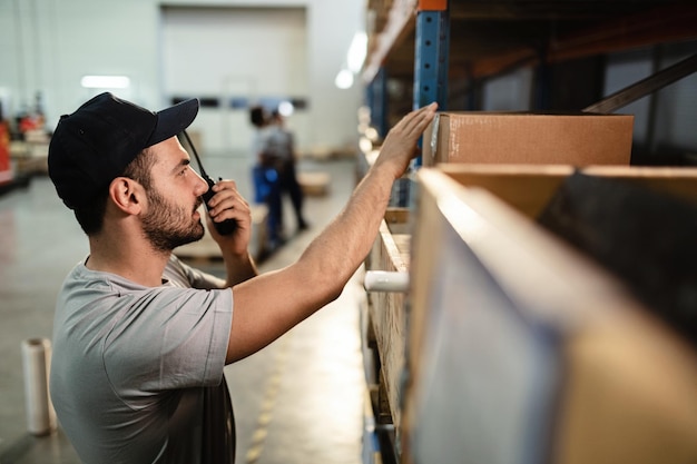 Jeune travailleur masculin vérifiant les colis sur une étagère tout en utilisant le talkie-walkie dans l'entrepôt de distribution