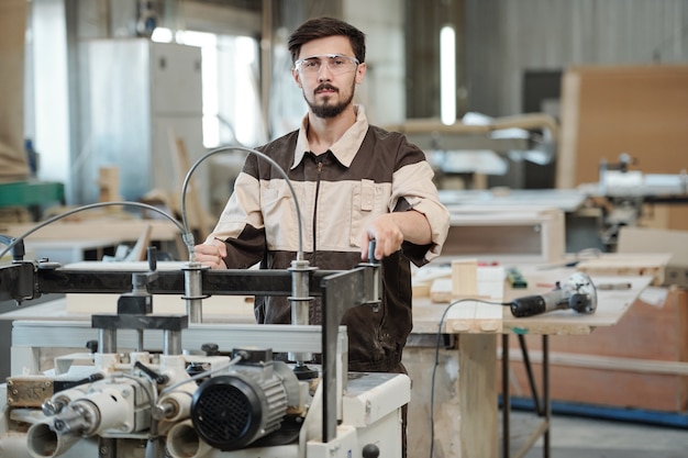 Jeune travailleur sérieux de l'usine de meubles vous regarde en se tenant debout par un établi